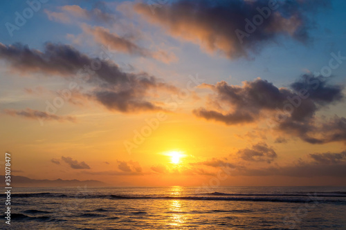 The sun rises over the ocean with backlit clouds. Beautiful sunrise at Pantai Pabean Ketewel Beach on the east coast of Bali. © EVISUAL