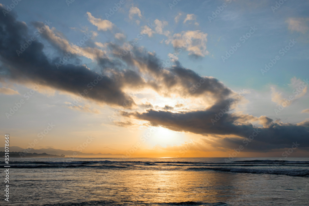 The sun rises over the ocean with backlit clouds. Beautiful sunrise at Pantai Pabean Ketewel Beach on the east coast of Bali.