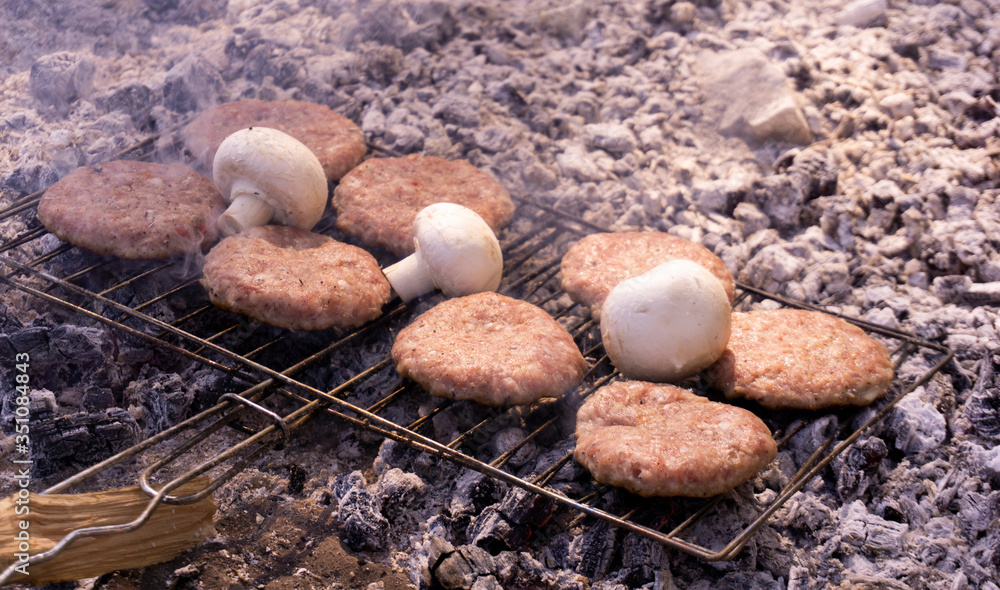 Beef BBQ Cooking of burger meat and mushroom. Iron barbeque grill pan with fire and food. Hamburgers outdoors barbecuing on coals. Closeup beef for burgers on cast iron grill. Juicy fat food grilled. Beef BBQ Cooking of burger meat and mushroom. Iron barbeque grill pan with fire and food. Hamburgers outdoors barbecuing on coals. Closeup beef for burgers on cast iron grill. Juicy fat food grilled.