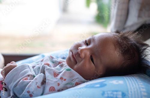 The adorable cute newborn baby infant with big black eyes lying on the bed with the view of garden on the window background