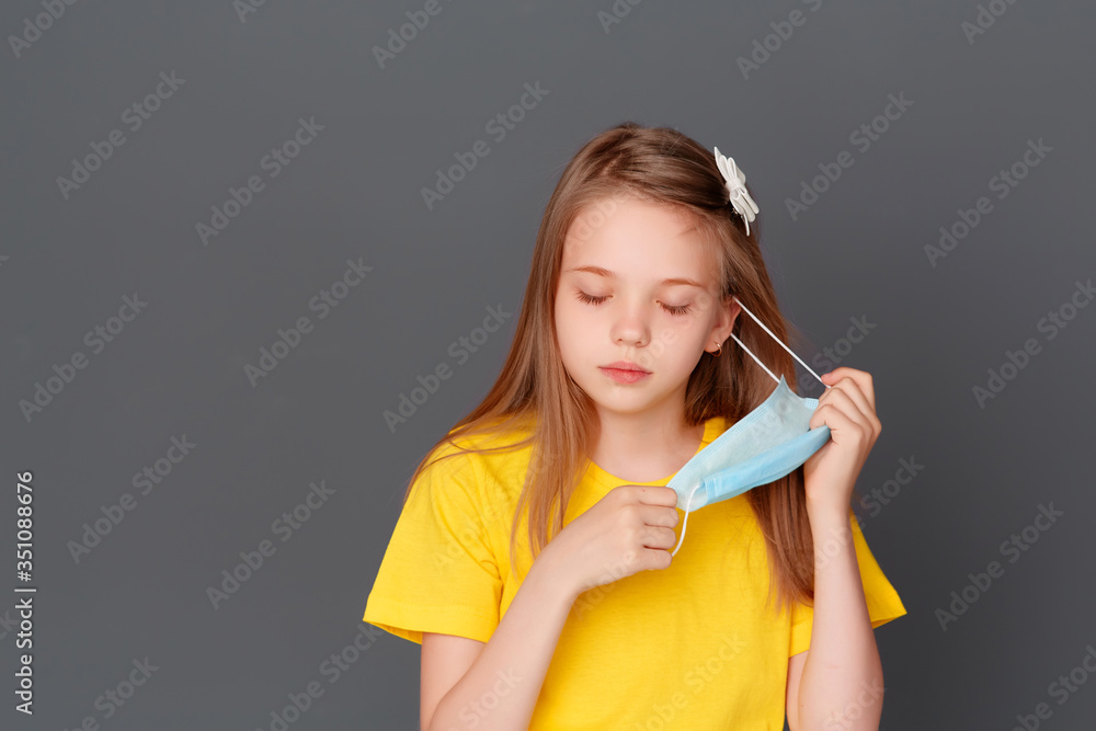 A little girl stands with a mask in her hand on a gray background, annoyed by the mask, she wants to take it off A little girl stands with a mask in her hand on a gray background, annoyed by the mask, she wants to take it off