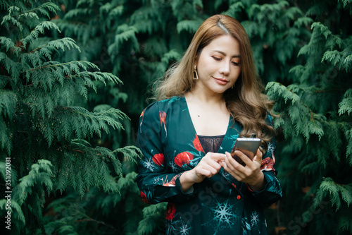 Beautiful asian woman in green dress using cellphone, mibile phone or smartphone pose in the park with green leaves. photo