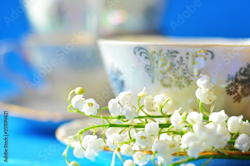 Part porcelain pattern teacup with golden handle and bunch white flower of lily of the valley on saucer close up on blue background with blurred heap teacups
