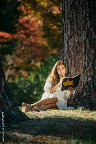 Beautiful asian woman in white dress sit under the tree writing and thinking in the park with autumn leaves.