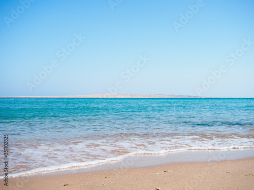 Red Sea photographed from beach, long view. Sea foam on brown sand. Blue sky is clear. Egypt in february, nature background. Selective soft focus. Blurred background