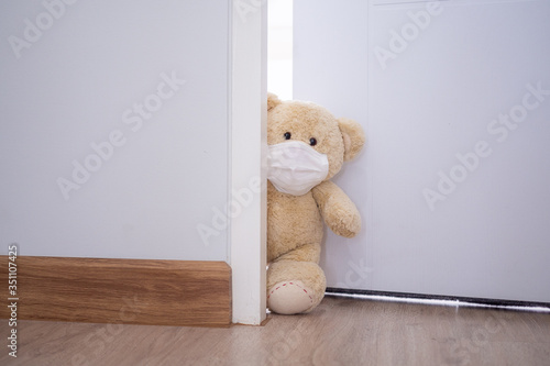 The teddy bear is wearing a mask to prevent the spread of the virus. The teddy bear stood behind the door inside the house. Children's concept
