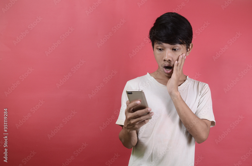 The face of Asian man is shocked what he sees in the smartphone. Asian men wearing white t shirts isolated on a red background The face of Asian man is shocked what he sees in the smartphone. Asian men wearing white t shirts isolated on a red background