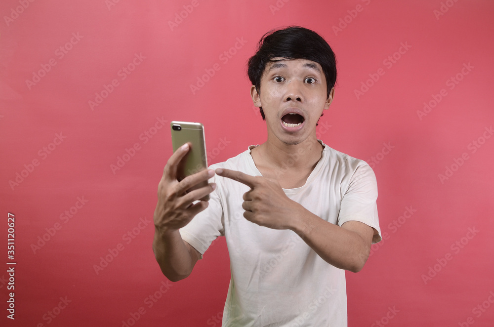 The face of Asian man is shocked what he sees in the smartphone. Asian men wearing white t shirts isolated on a red background The face of Asian man is shocked what he sees in the smartphone. Asian men wearing white t shirts isolated on a red background