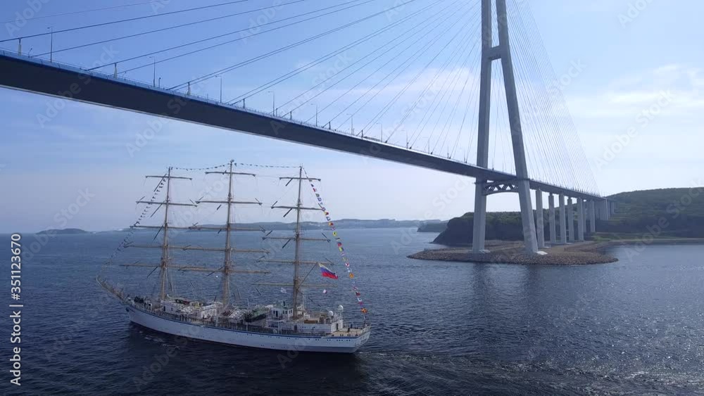 Picturesque old large ship sailing frigate floats under Russian unique cable bridge. Flag wind. Tourist landmark. Vladivostok Russia cinematic event. Summer sunny. Aerial flight together with ship Picturesque old large ship sailing frigate floats under Russian unique cable bridge. Flag wind. Tourist landmark. Vladivostok Russia cinematic event. Summer sunny. Aerial flight together with ship