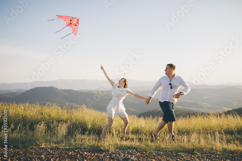 Running with flying kite have fun on countryside. Boy and girl running on a hill with a flying kite against a blue sky and sun rays.