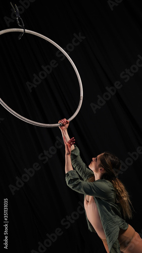 Beautiful graceful girl, aerialist. professional circus acrobat on an aerial hoop. performer holding a ring in his hands, black background. black curtain backstage with pleats, rehearsal, grace