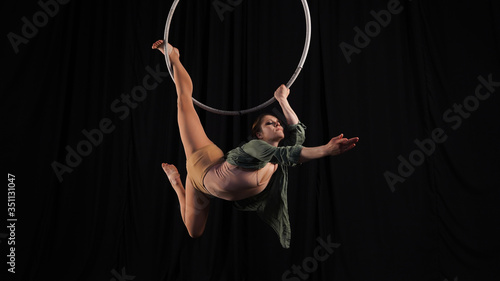 Aerial gymnast on the hoop. Young graceful girl rubber, flexibility in the air ring. Danger, risk. Female circus performer on a black background in the studio.