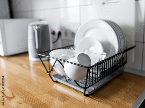 Dish rack holds many dishes and cups against wooden countertop, white wall tiles, sink and faucet. Budget and lightweight antimicrobial dish drainer with drain board at modern scandinavian kitchen photo