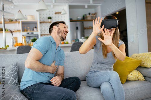 Smiling young woman using VR headset at home on couch. Woman and her husband enjoying virtual reality at her apartment.