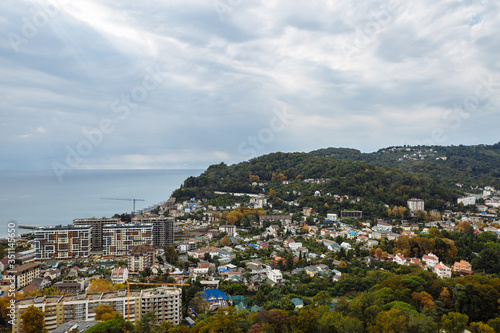 View of a small resort town near the city of Sochi. The photo shows the Black Sea, small hotels, residential buildings, mountains, green trees and blue sky.