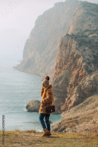 girl in a brown fur coat, in a knitted hat with a boumbon, with flying hair on a cliff, against the backdrop of rocks, mountains and the sea. cloudy sky, clouds.
