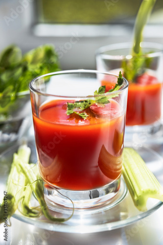 Two glasses of tomato juice with parsley and celery decorations, stand near the window, morning sunlight shines, shallow depth of field, selective focus. Natural drinks concept.