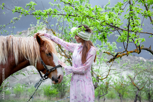 Beautiful woman dressed pink dress and fashion headdress, walking outdoor near horse paddock in the ranch. Friendship lifestyle of woman with wild animal in the nature on the garden