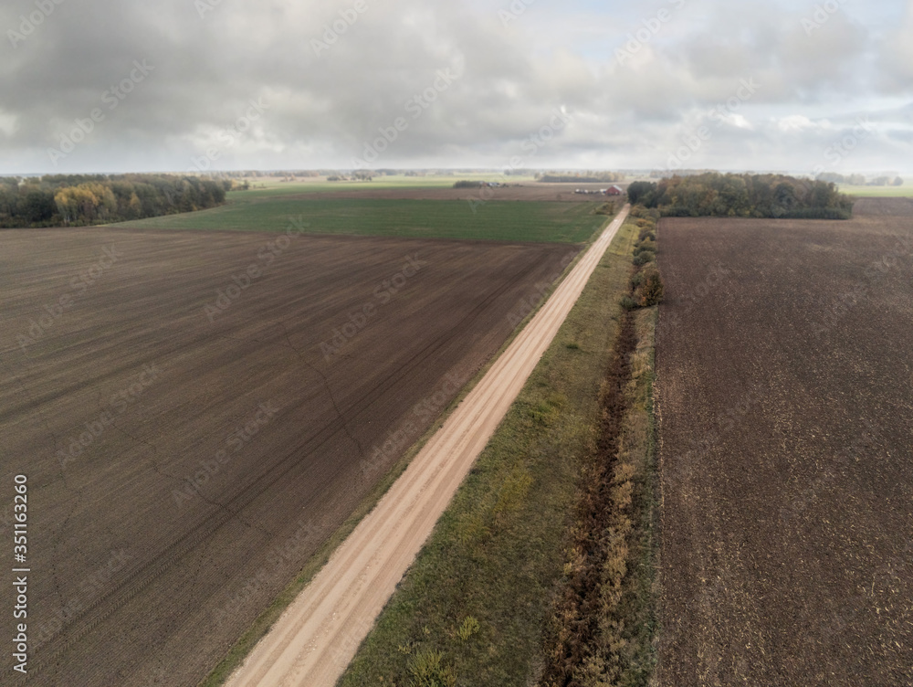 Aerial view on a freshly ploughed field, cloudy sky. Small spot with trees and small country road.