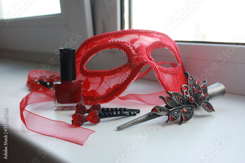 Composition of red vintage carnival mask, nail polish, metal hair clips with rhinestones, pomegranate beads made of natural stone on a white background on the windowsill. photo