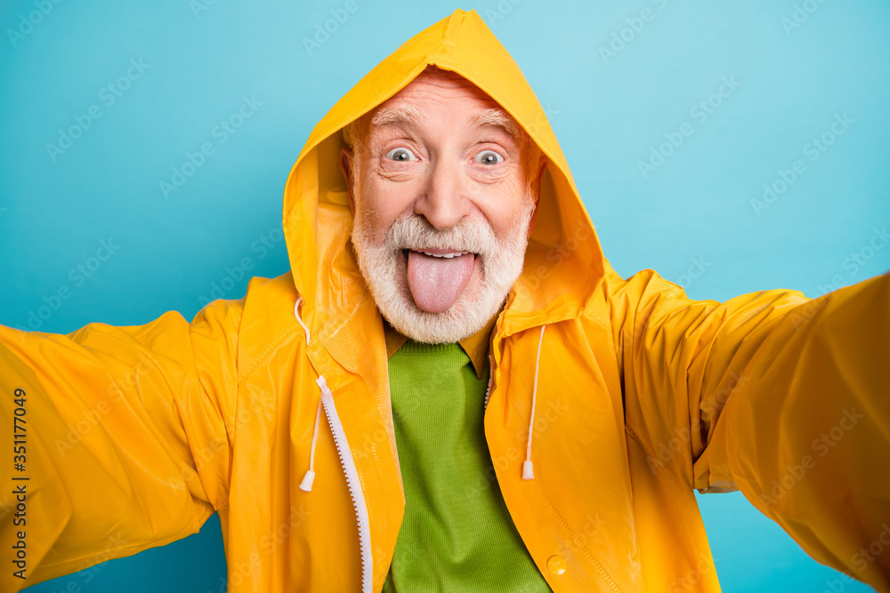 Self-portrait of his he nice cheerful cheery crazy foolish grey-haired man wearing yellow topcoat having fun showing tongue out isolated on bright vivid shine vibrant blue color background
