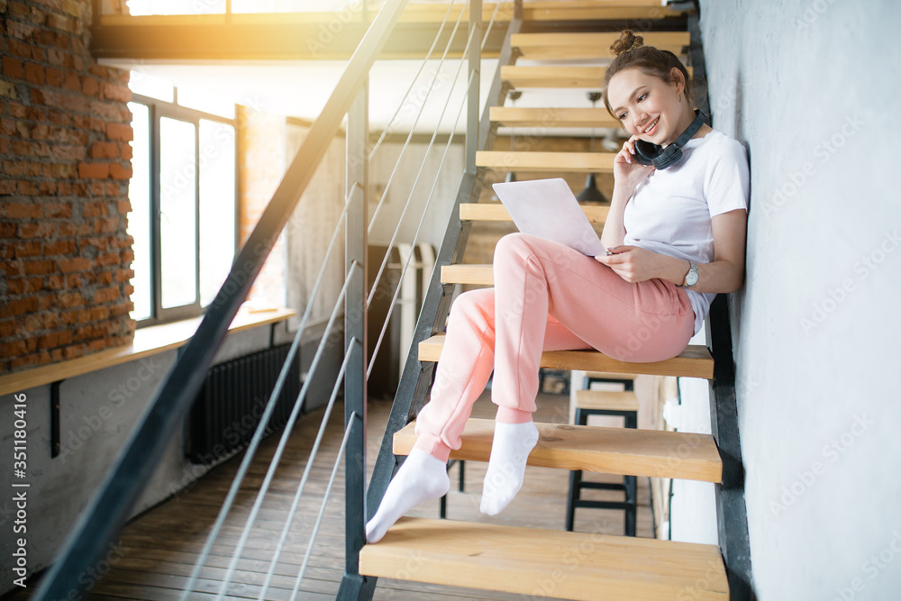 With a laptop on her lap, the girl is sitting in the house on the stairs. student in headphones communicates online with friends With a laptop on her lap, the girl is sitting in the house on the stairs. student in headphones communicates online with friends