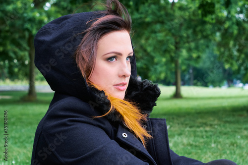 Pensive young woman with gray eyes and an ombre hairstyle, glancing thoughtful into the distance while sitting in a green park, wearing a black hooded fur lined wool coat.