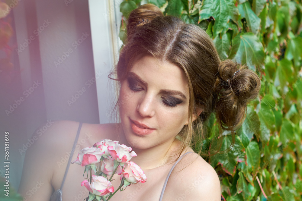 Young natural woman looking at a white and red roses flower bouquet while standing at her garden window blending in between violett dreamy foliage with a soft pastel bokeh.