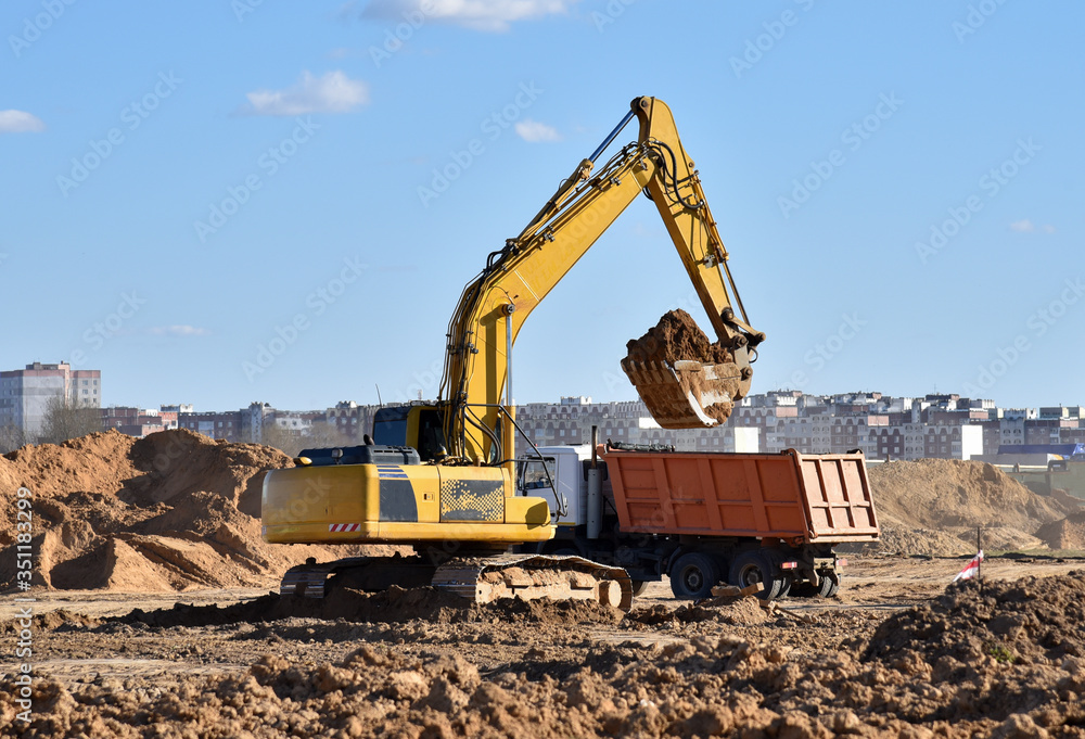 Excavator load the sand to the heavy dump truck on construction site. Backhoe digs the ground for the foundation and for laying sewer pipes. Renovation program. Buildings industry background Excavator load the sand to the heavy dump truck on construction site. Backhoe digs the ground for the foundation and for laying sewer pipes. Renovation program. Buildings industry background