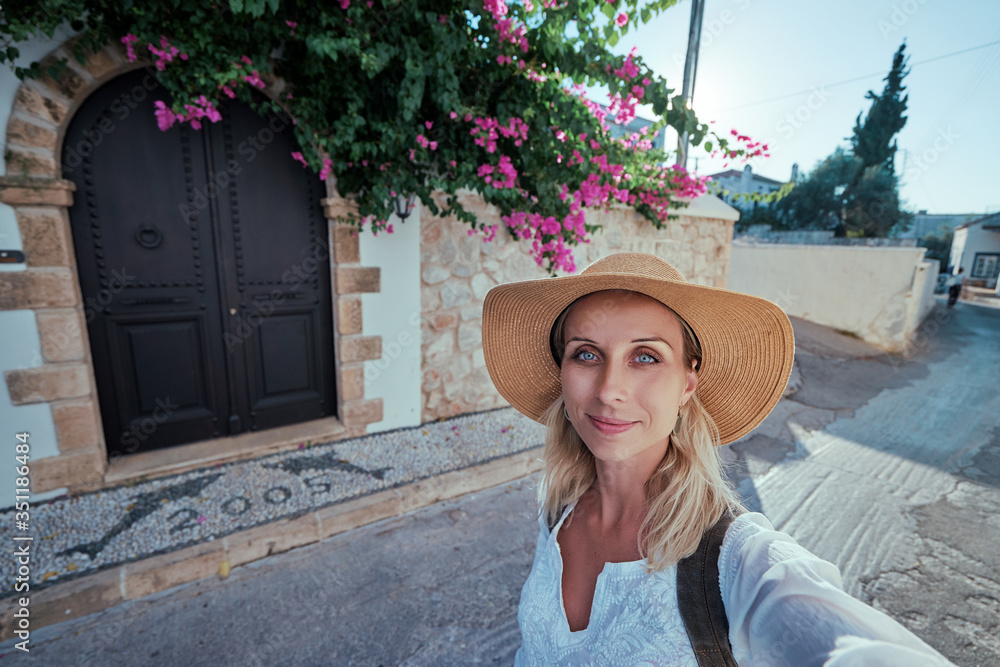 Travel by Greece, Young woman in hat with rucksack taking selfie near traditional greek house.