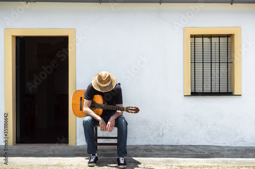 Young man sitting in front of the facade of his rural house in the countryside takes a break from playing guitar on sunny day. Rural lifestyle.