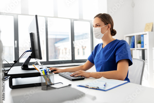 medicine, technology and healthcare concept - female doctor or nurse wearing face protective medical mask for protection from virus disease with computer working at hospital