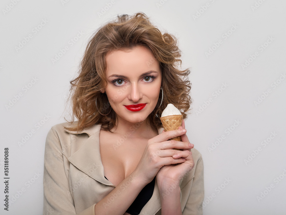 Sexy woman with red lips curly hair in short coat and black bra playfully posing with delicious ice cream on white background looking into camera Sexy woman with red lips curly hair in short coat and black bra playfully posing with delicious ice cream on white background looking into camera