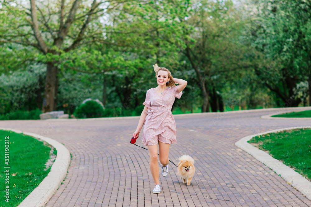 Fototapeta premium Portrait of a young glamorous female wearing pink dress holding her cute pomeranian spitz on hands. . Concept friendship between dogs and humans.