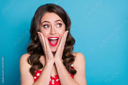 Closeup photo of pretty funny shocked wavy lady arms on cheeks open mouth excited overjoyed expression look side empty space wear red dotted dress singlet isolated blue color background