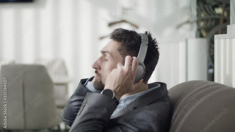 Young brunette man putting on headphones and leaning back in armchair. Side view portrait of handsome Caucasian businessman resting in VIP departure area of the airport. Lifestyle, relaxation, tourism Young brunette man putting on headphones and leaning back in armchair. Side view portrait of handsome Caucasian businessman resting in VIP departure area of the airport. Lifestyle, relaxation, tourism