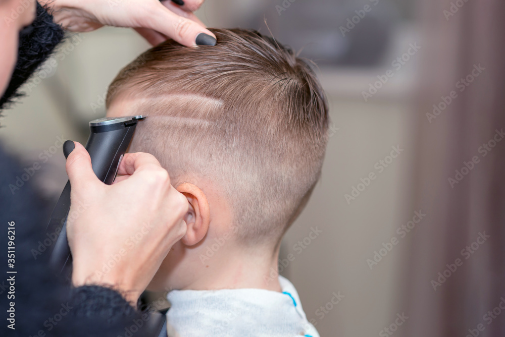 Fototapeta premium Hairdresser makes a stylish hairstyle. The woman is standing and making haircut for blonde boy. Close up of hands.