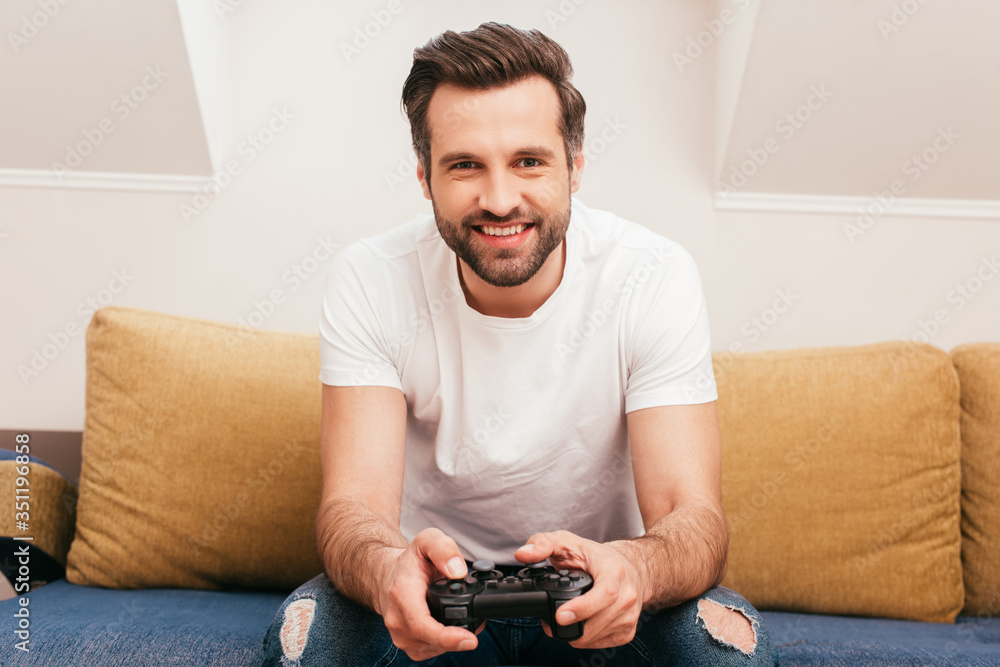 KYIV, UKRAINE - APRIL 14, 2020: Handsome man smiling at camera and holding joystick on sofa at home KYIV, UKRAINE - APRIL 14, 2020: Handsome man smiling at camera and holding joystick on sofa at home