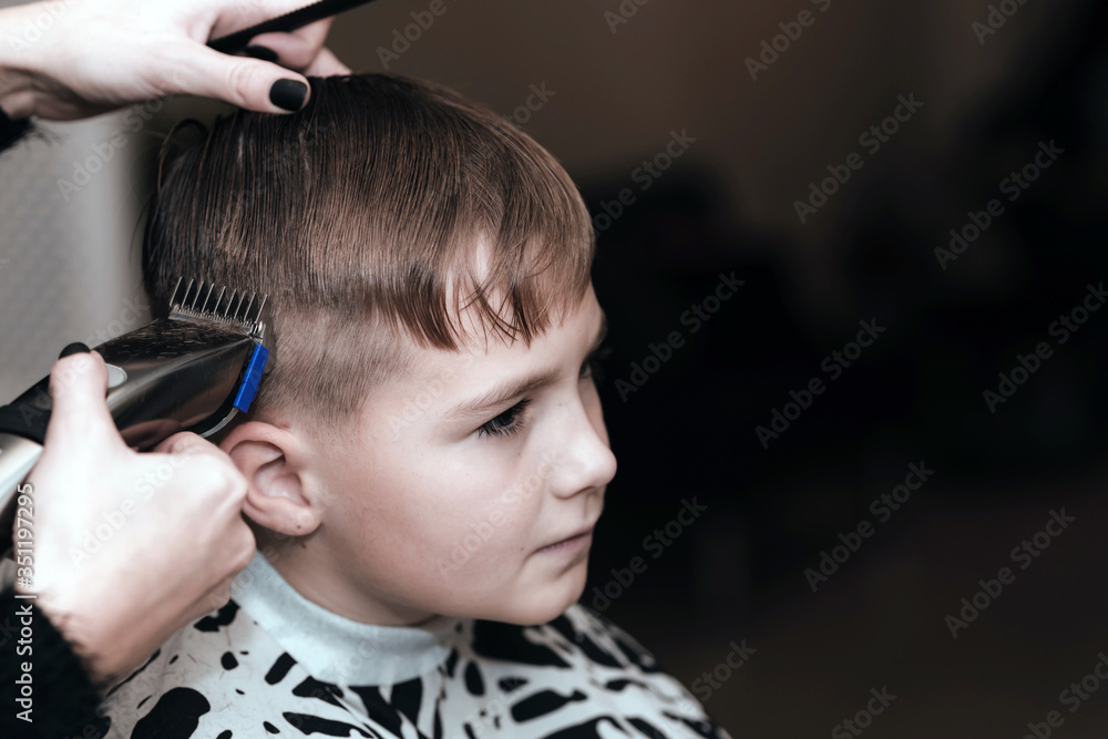 Fototapeta premium Toned image.Hairdresser makes a stylish hairstyle. The woman is standing and making haircut for blonde boy. Close up of hands.
