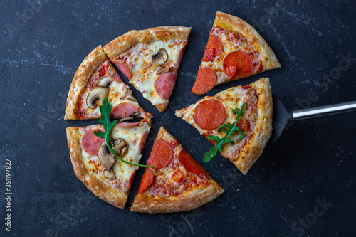A fresh prepared pizza with salami, mushrooms, ham and cheese on a dark background. Italian traditional lunch or dinner. Fast food and street food concept. Flat lay, top view, close up