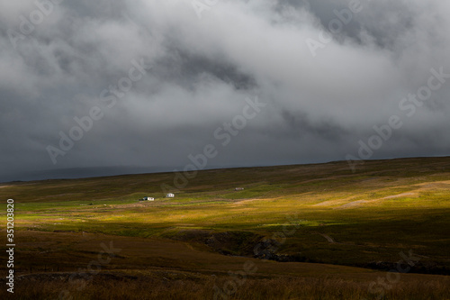 landscape in iceland. There is a lonely house in the middle of a green meadow. the sky is full of clouds and we can see torrents of water falling in the middle of the sun's rays