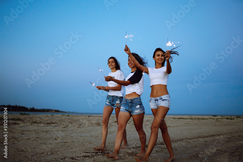 Three beautiful girls celebrating, holding sparklers on the beach at night. Young teenagers enjoying on beach holiday. Summer holidays, vacation, relax and lifestyle concept.