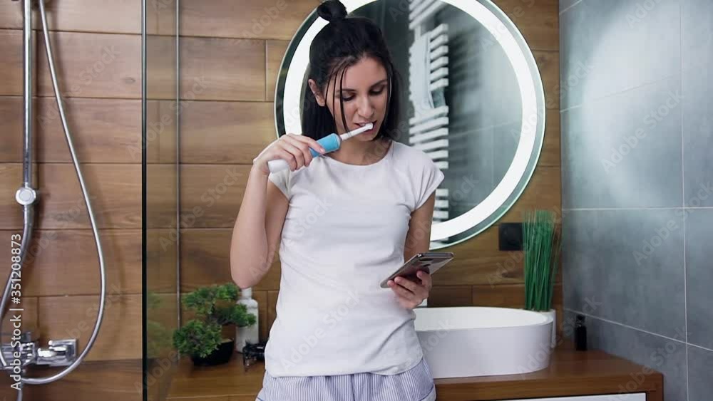 Front view of attractive smiling young brunette which brushing her teeth and watching video on her phone Front view of attractive smiling young brunette which brushing her teeth and watching video on her phone