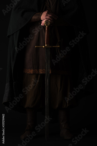 cropped view of medieval Scottish man in mantel with sword in dark isolated on black