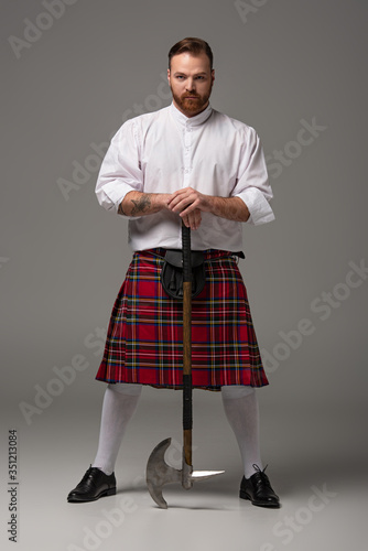 serious Scottish redhead man in red kilt with battle axe on grey background
