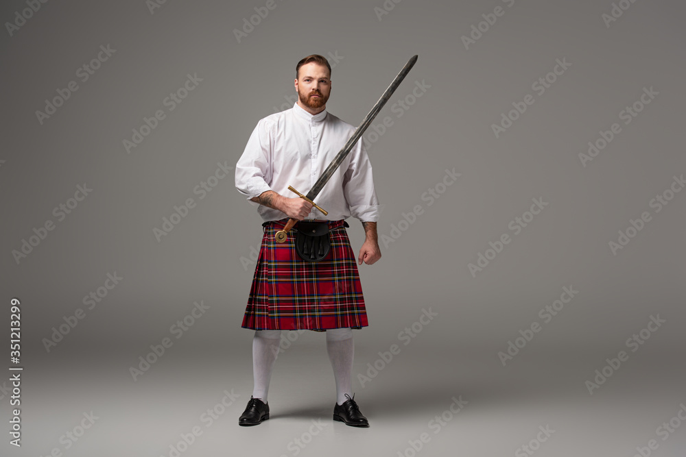 Scottish redhead man in red kilt with sword on grey background Scottish redhead man in red kilt with sword on grey background