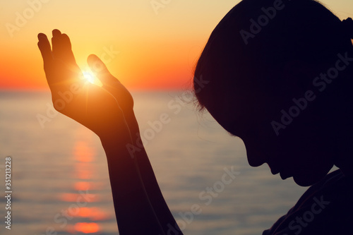 silhouette of young woman raising hands praying at sunset or sunrise light, yoga practice, religion, freedom and spirituality concept