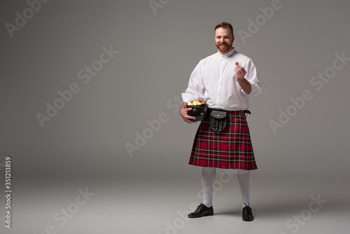 smiling Scottish redhead man in red kilt holding potty with gold coins on grey background