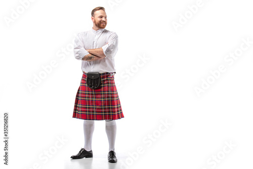 smiling Scottish redhead man in red kilt with crossed arms on white background