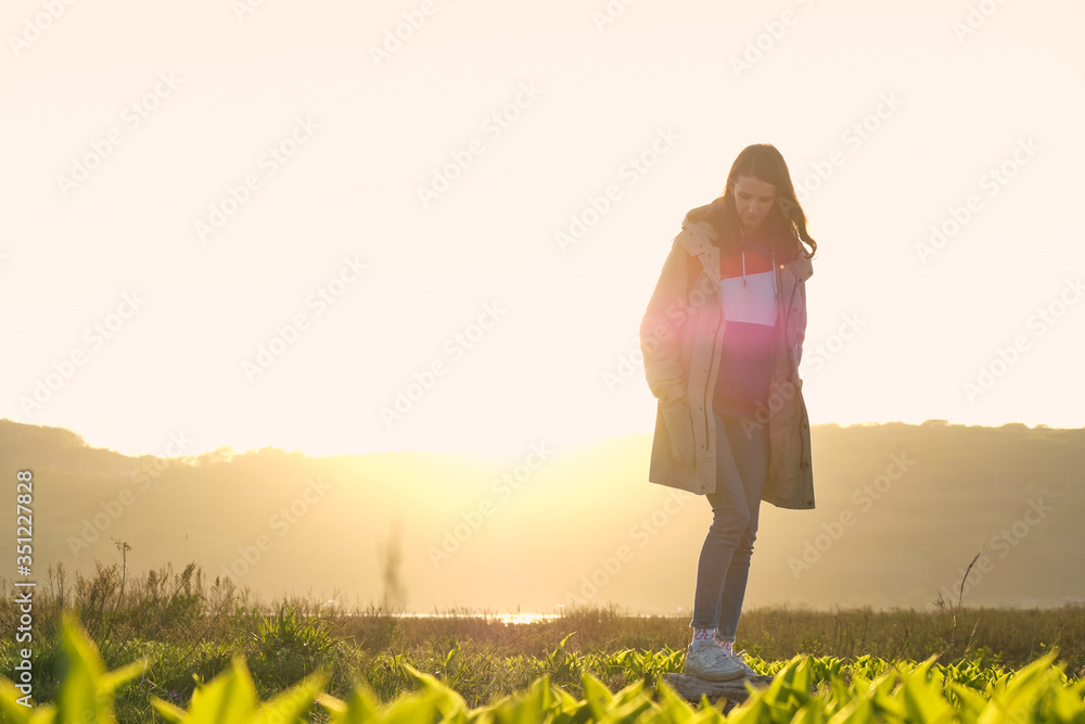 Fototapeta premium Close up young girl balancing on a wooden log in the middle of a green field in the sunset rays. Journey and outdoor concept.